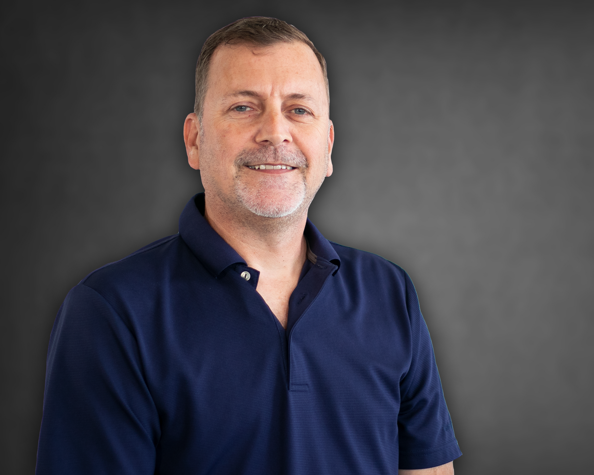 A man with a beard is wearing a blue polo shirt and smiling.