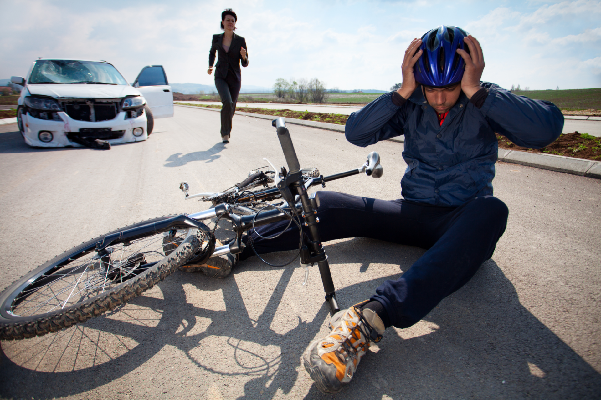 Man on ground with bike after car crash; woman runs toward them, damaged car in background.