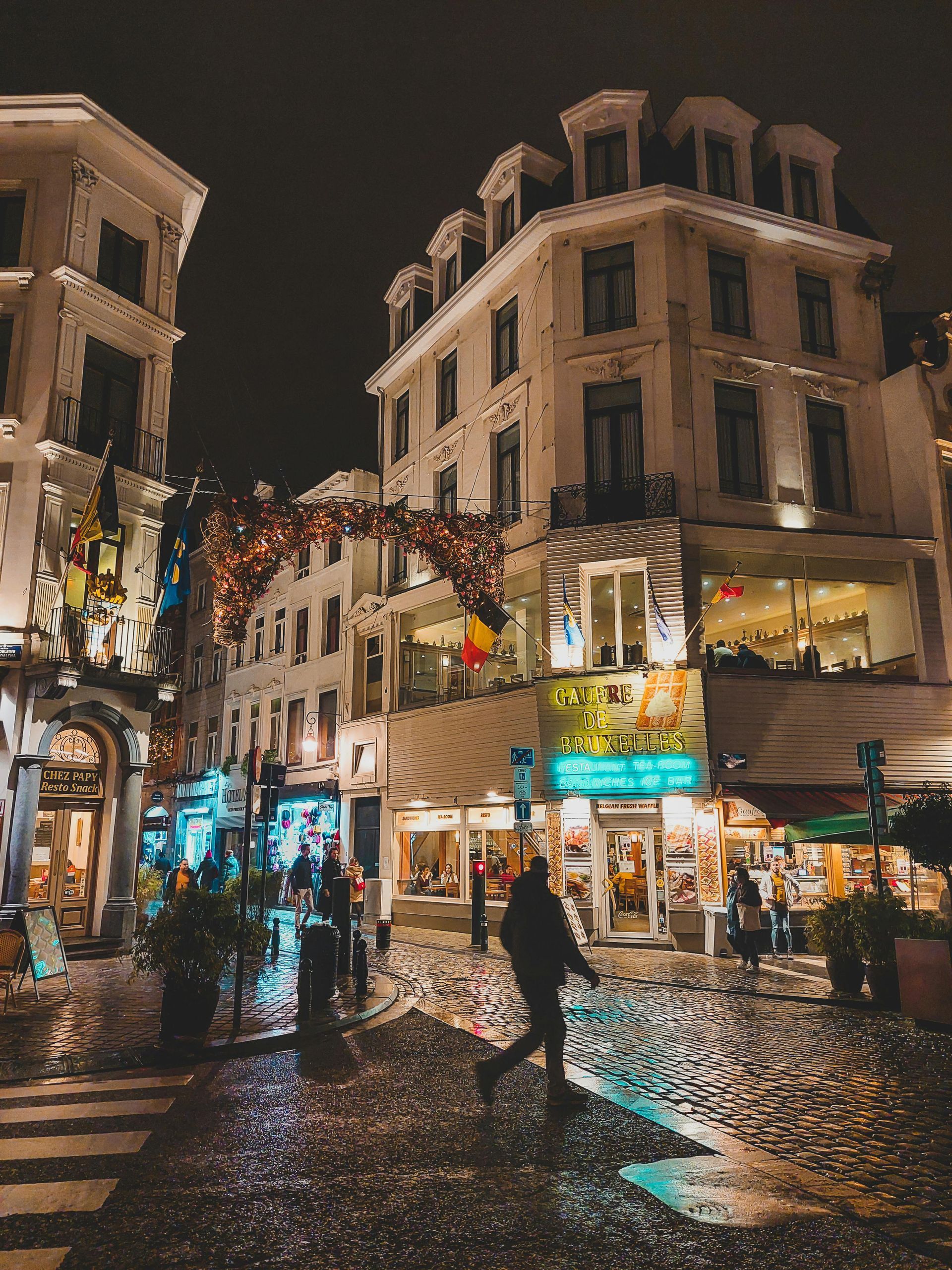 Cobblestone street at night, lined with illuminated buildings and shops; a person walks by.