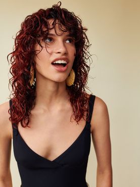 A woman with red curly hair is wearing a black tank top and earrings