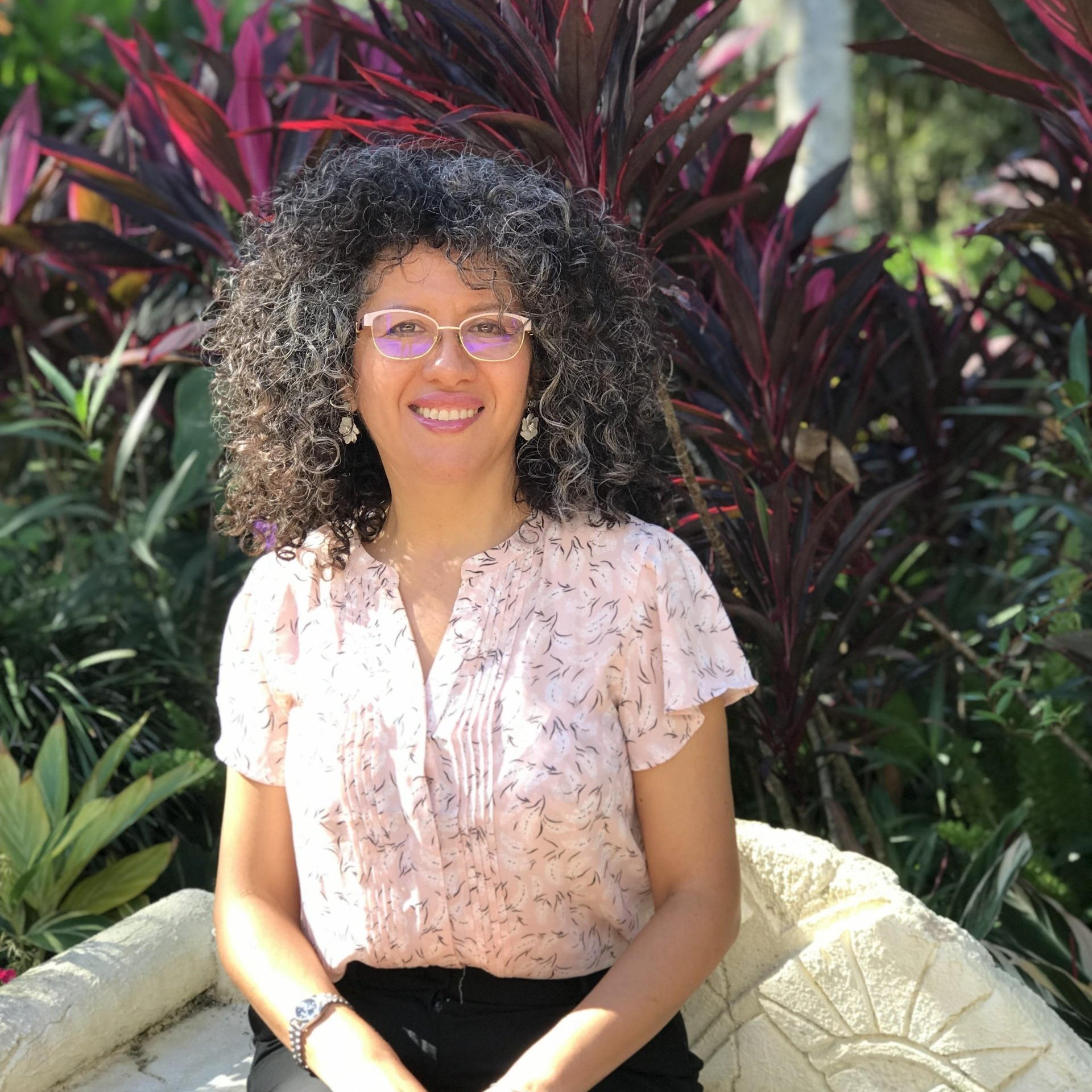 A woman with curly hair and glasses is sitting on a chair in front of plants.