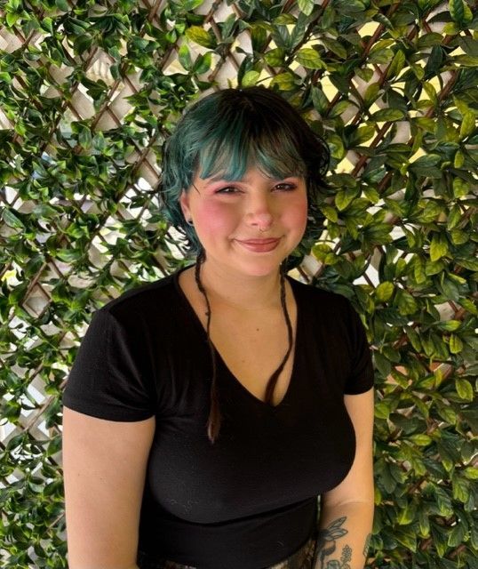 A woman with green hair and a black shirt is standing in front of a hedge.