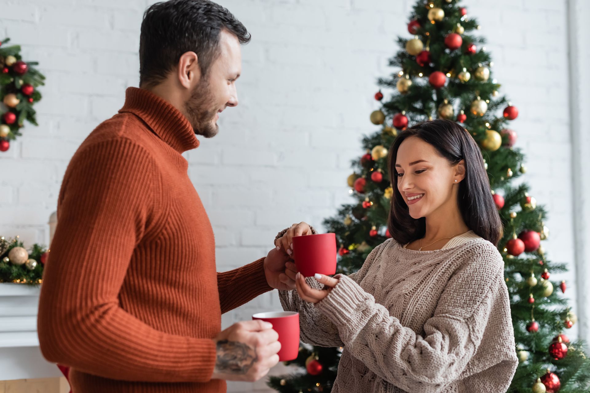 A man and a woman are holding cups of coffee in front of a christmas tree.