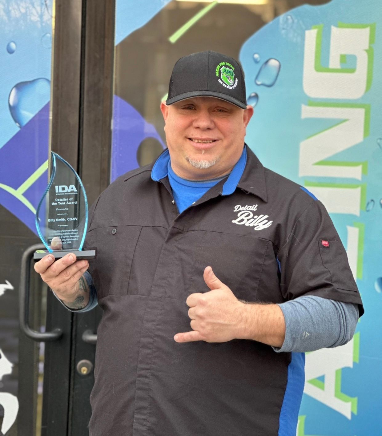 Man holding an award, thumbs up. Wearing a baseball cap and work shirt outside a business.