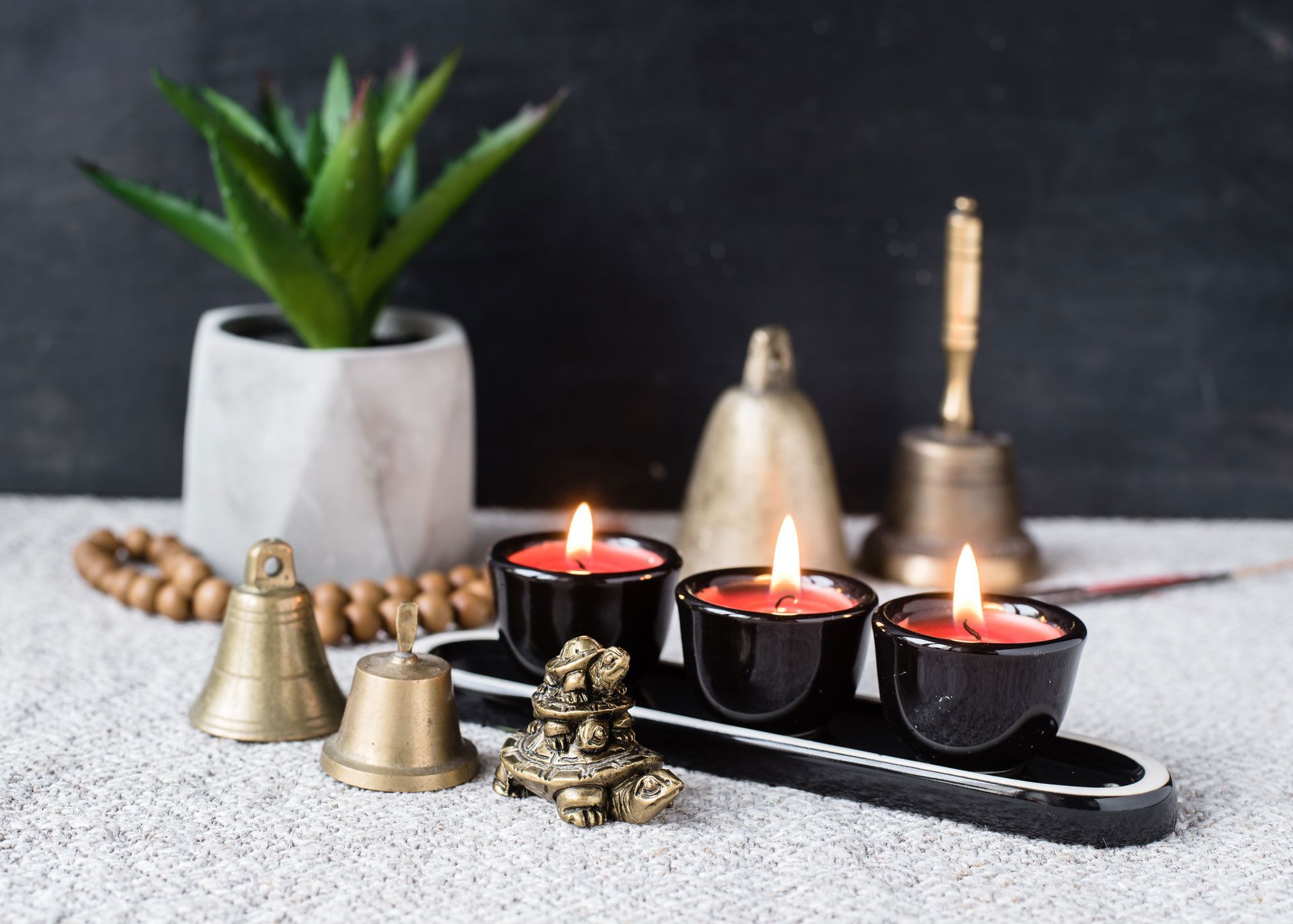 Candles burning in black holders, surrounded by bells, a plant, and beads on a textured surface.
