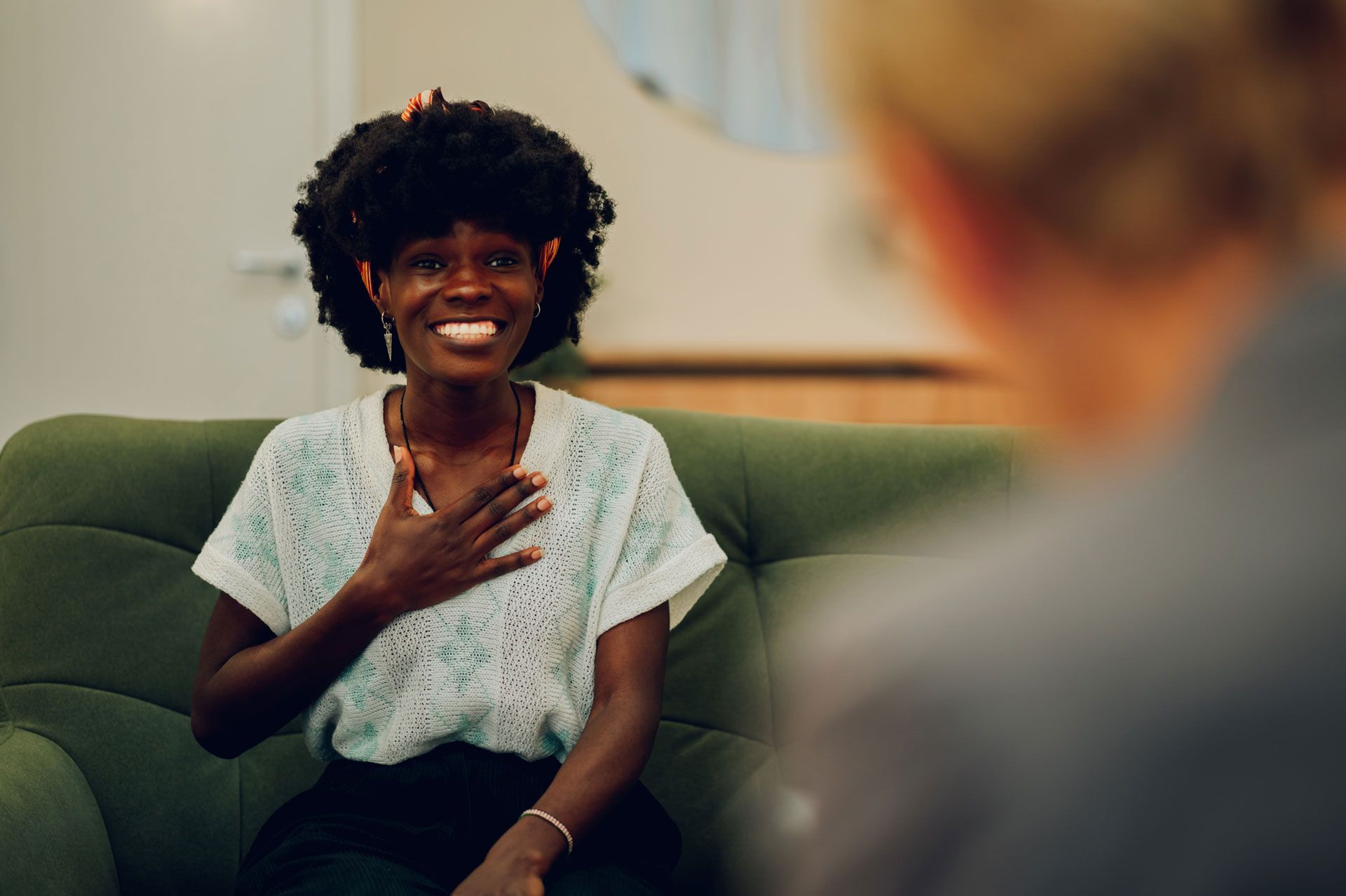 Woman smiles, placing her hand over her heart while sitting on a green couch, facing another person.