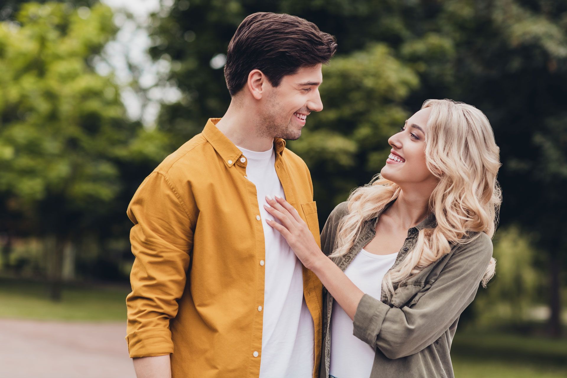 Couple smiling at each other in a park. Woman's hand on man's chest. Green trees in background.