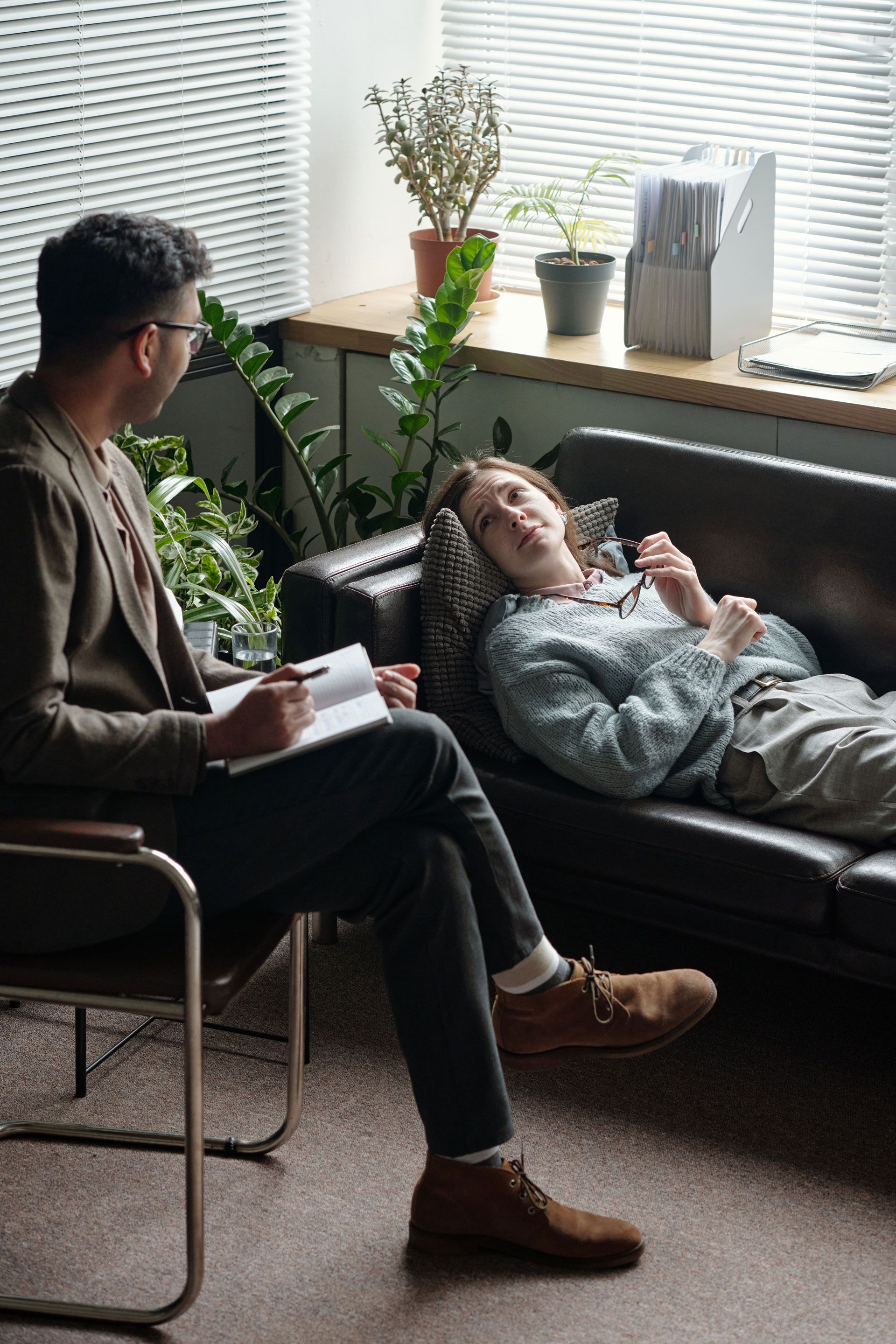 Man in a cardigan takes notes as person reclines on a couch. The setting is an office, plants in background.