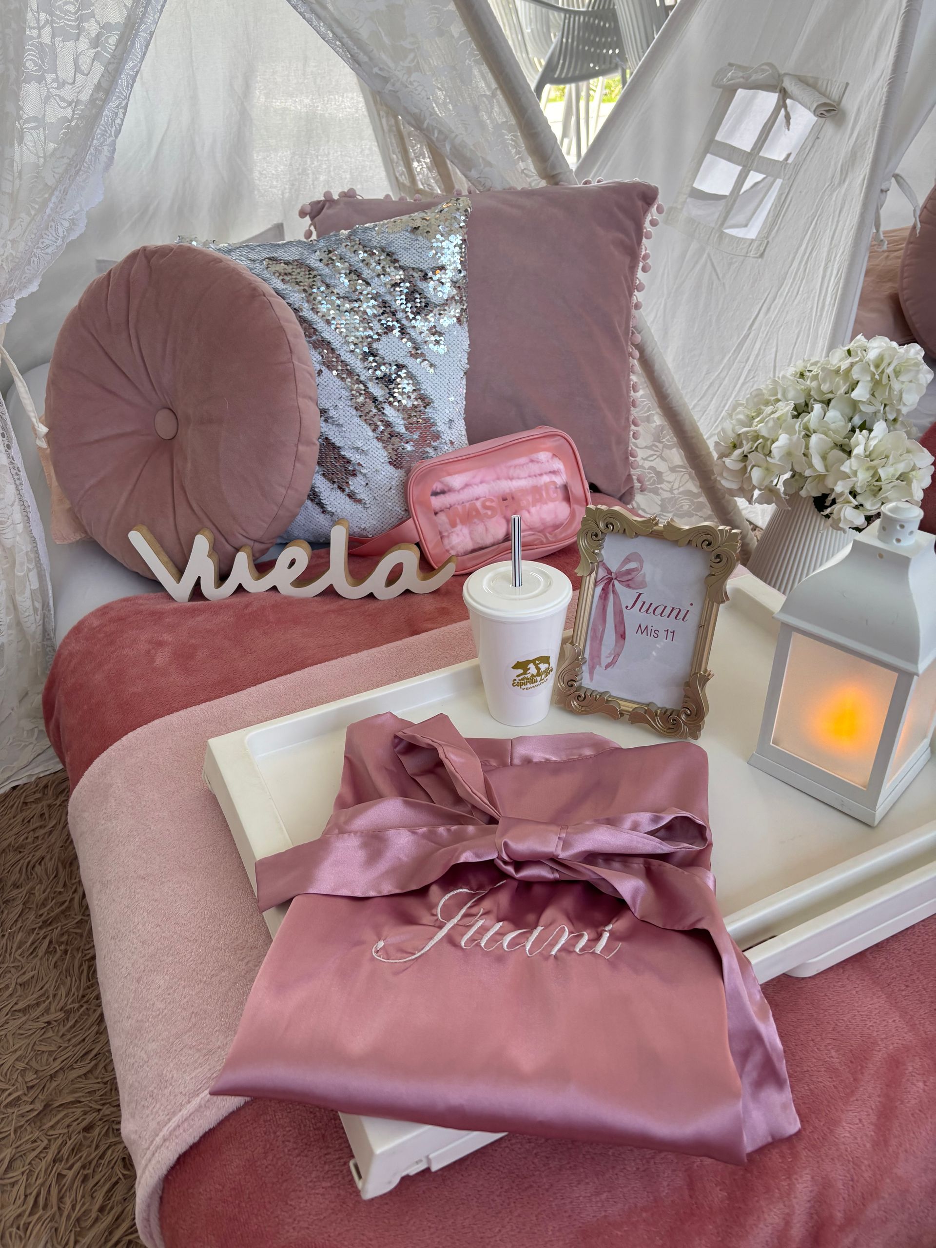 Configuración temática rosa con bata en bandeja, almohadas, arreglo floral y un marco.