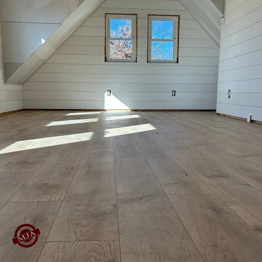 A room with light wood floors, white shiplap walls, and two windows reflecting blue sky, with bright light on the floor.