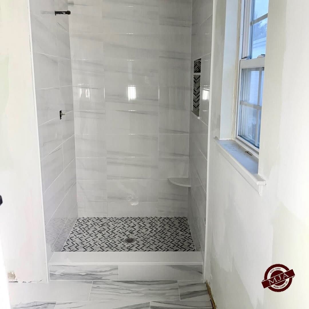 A tiled walk-in shower with a white marble-patterned wall, a dark patterned floor, a small built-in shelf, and a window.