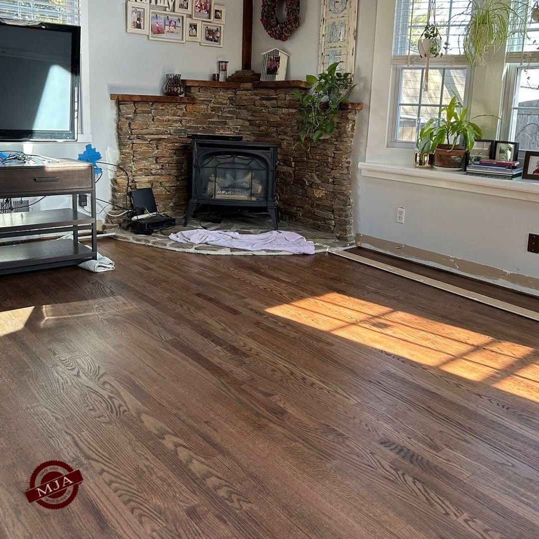 A living room with dark hardwood floors, a stone corner fireplace with a black stove, and a window with plants.