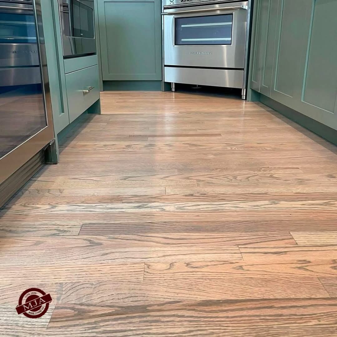 A view of light-toned hardwood flooring in a kitchen with sage green cabinets and a stainless steel oven.