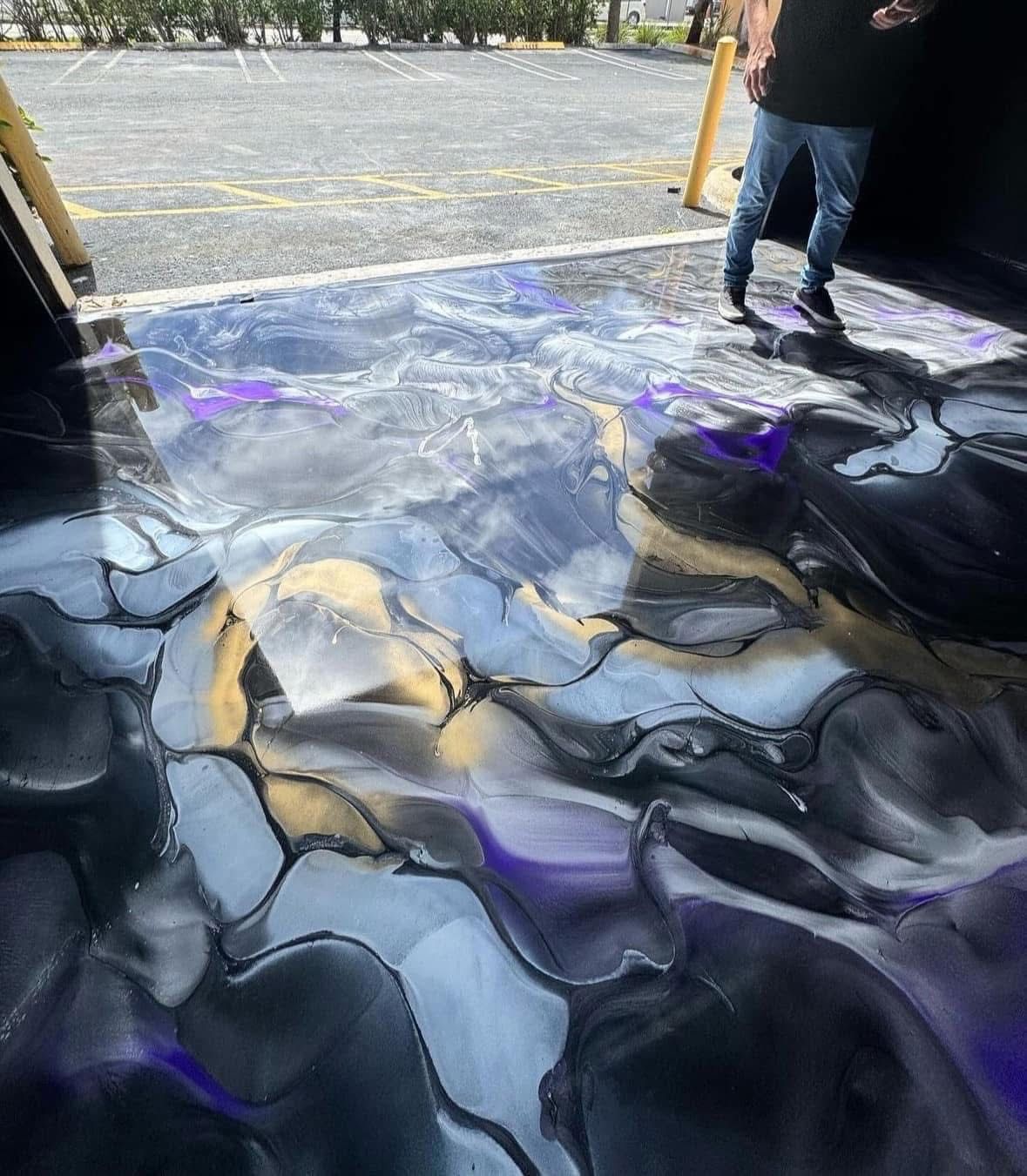A freshly poured epoxy floor with a swirling, marbled pattern of black, gray, purple, and gold near an open garage door.