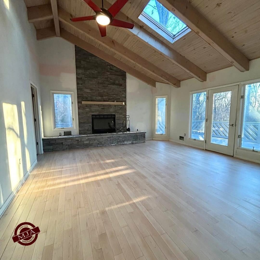 A spacious room with light hardwood floors, a vaulted wood-beam ceiling, stone fireplace, and floor-to-ceiling windows.
