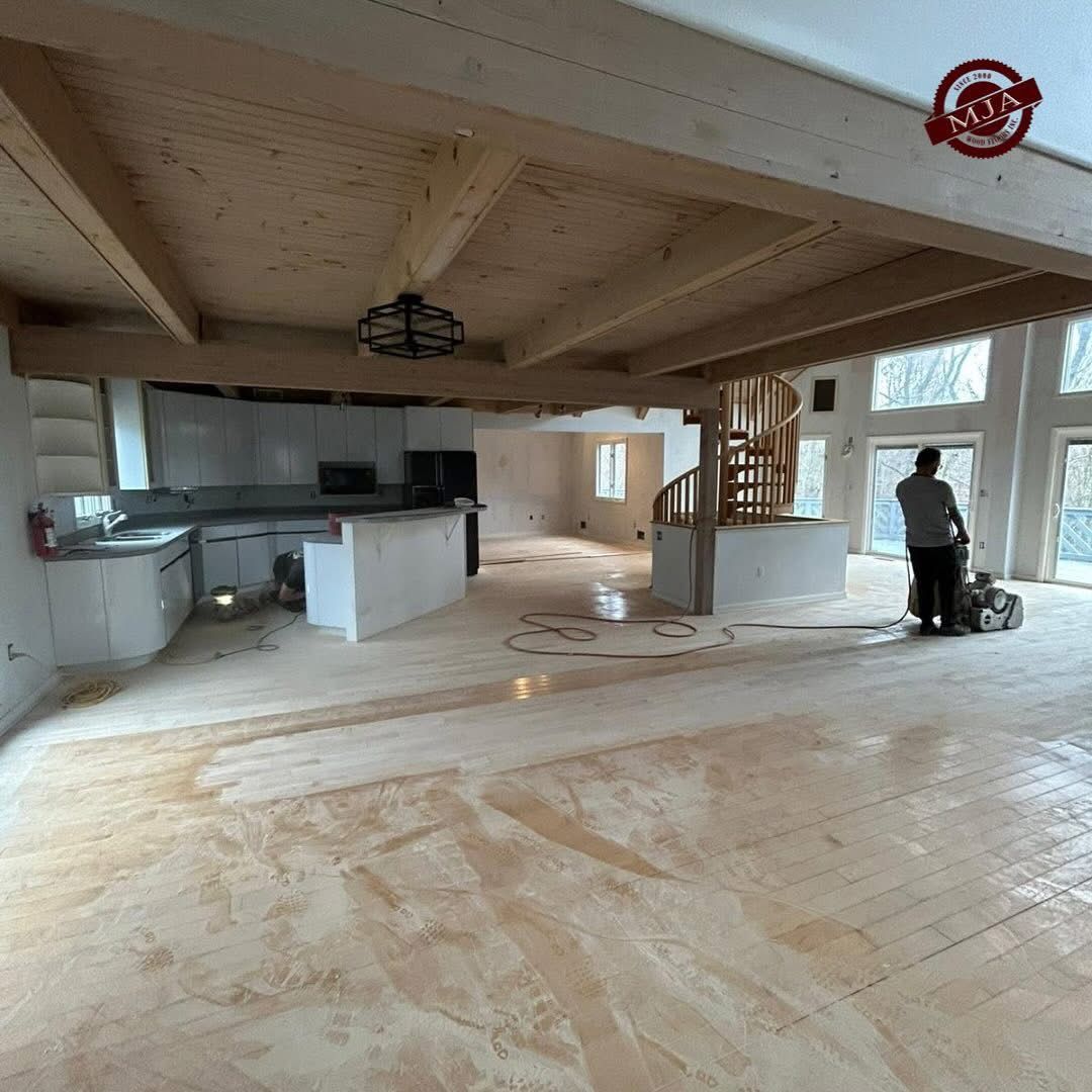 A person using a floor sander in a large, open-concept room with a spiral staircase, kitchen, and exposed wooden beams.