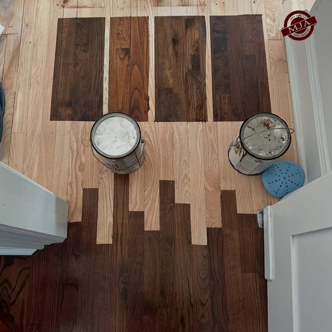 Hardwood floor samples in four different brown stains arranged on unfinished wood, with two paint cans nearby.