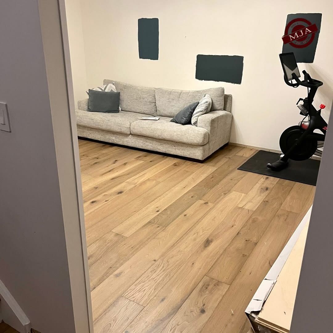 A light-filled room featuring a light gray couch, a stationary exercise bike on a black mat, and wood-tone flooring.