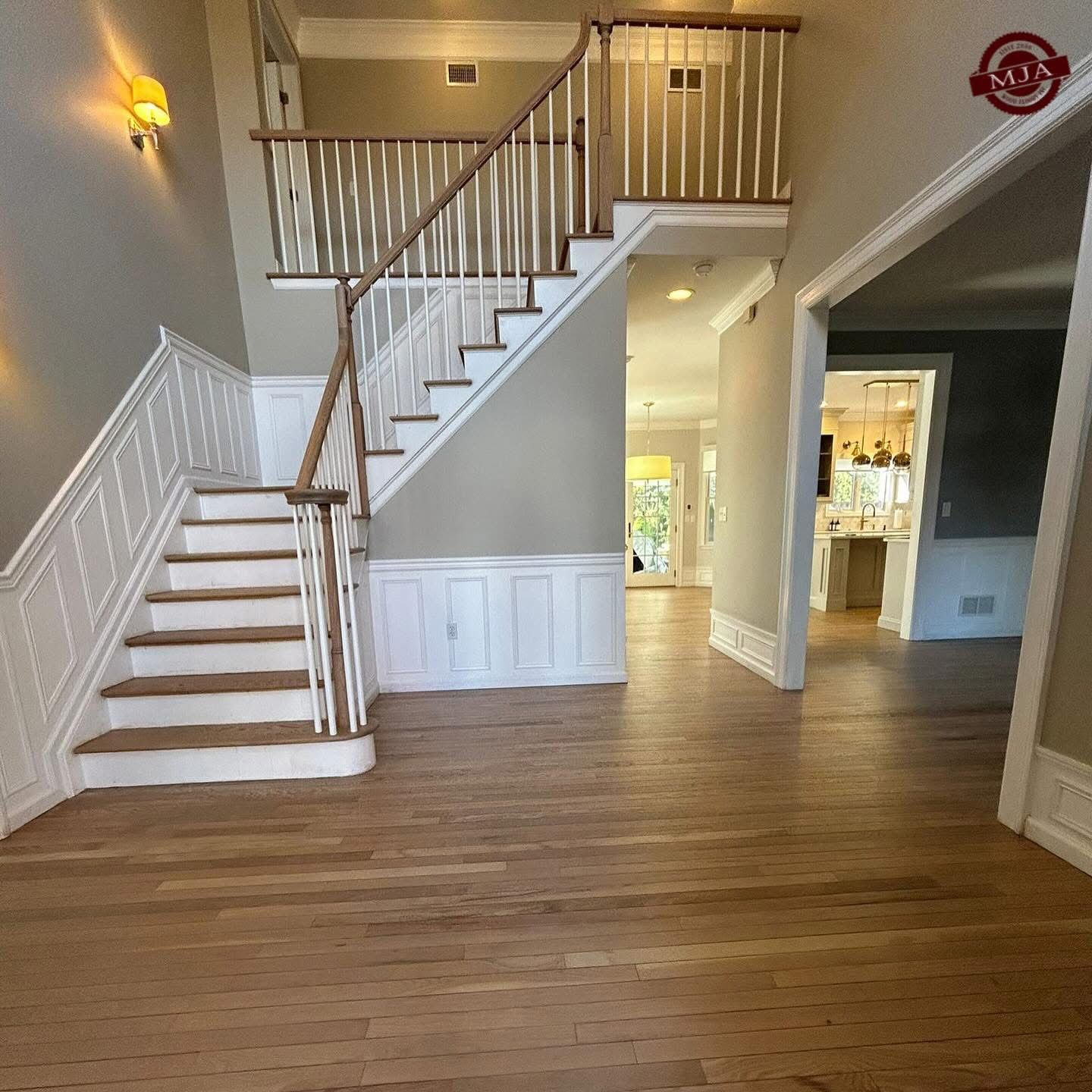 An interior view of an entryway with a wooden staircase, light hardwood floors, and a glimpse into a kitchen area.