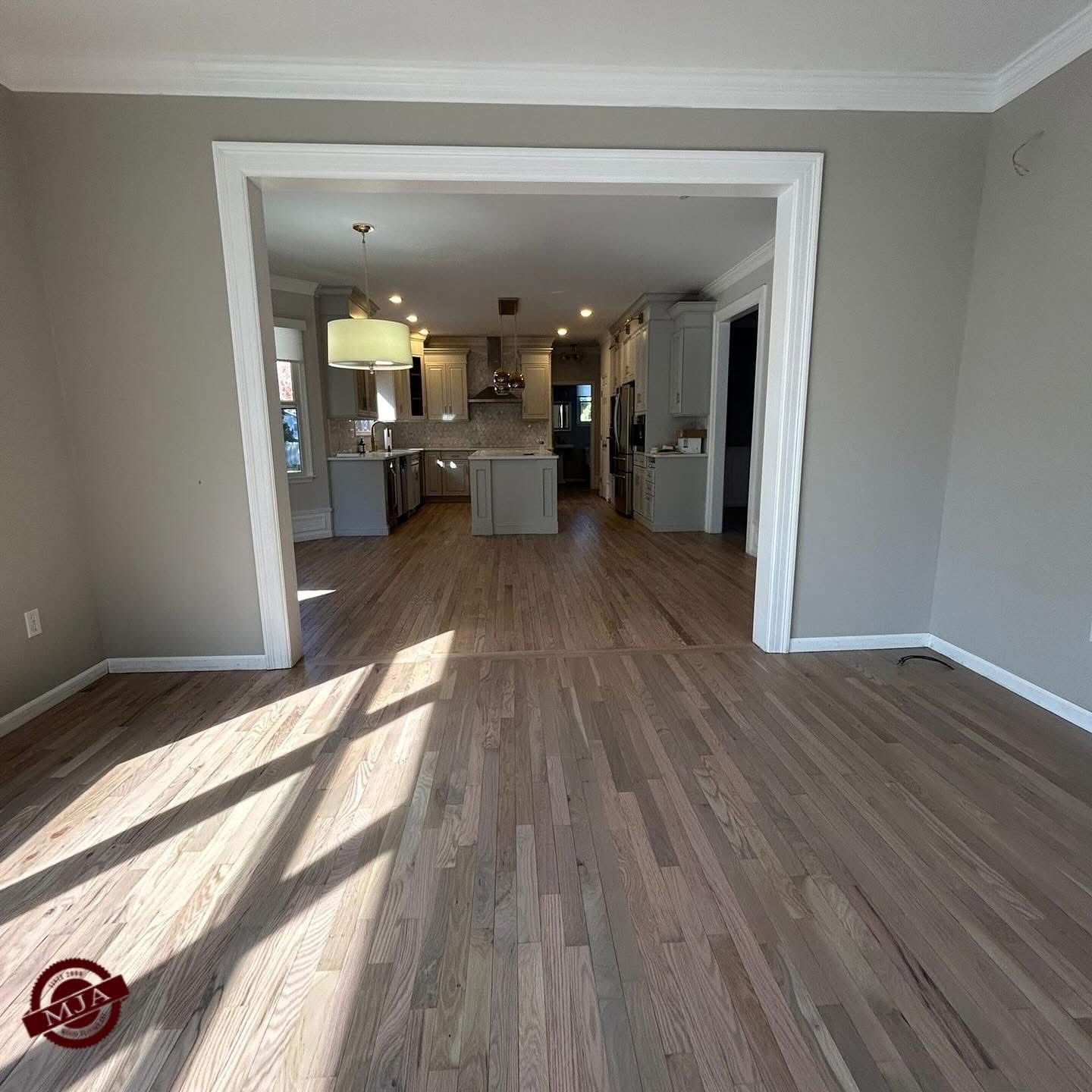 An open floor plan room features light hardwood floors and gray walls, looking through a doorway into a modern kitchen.