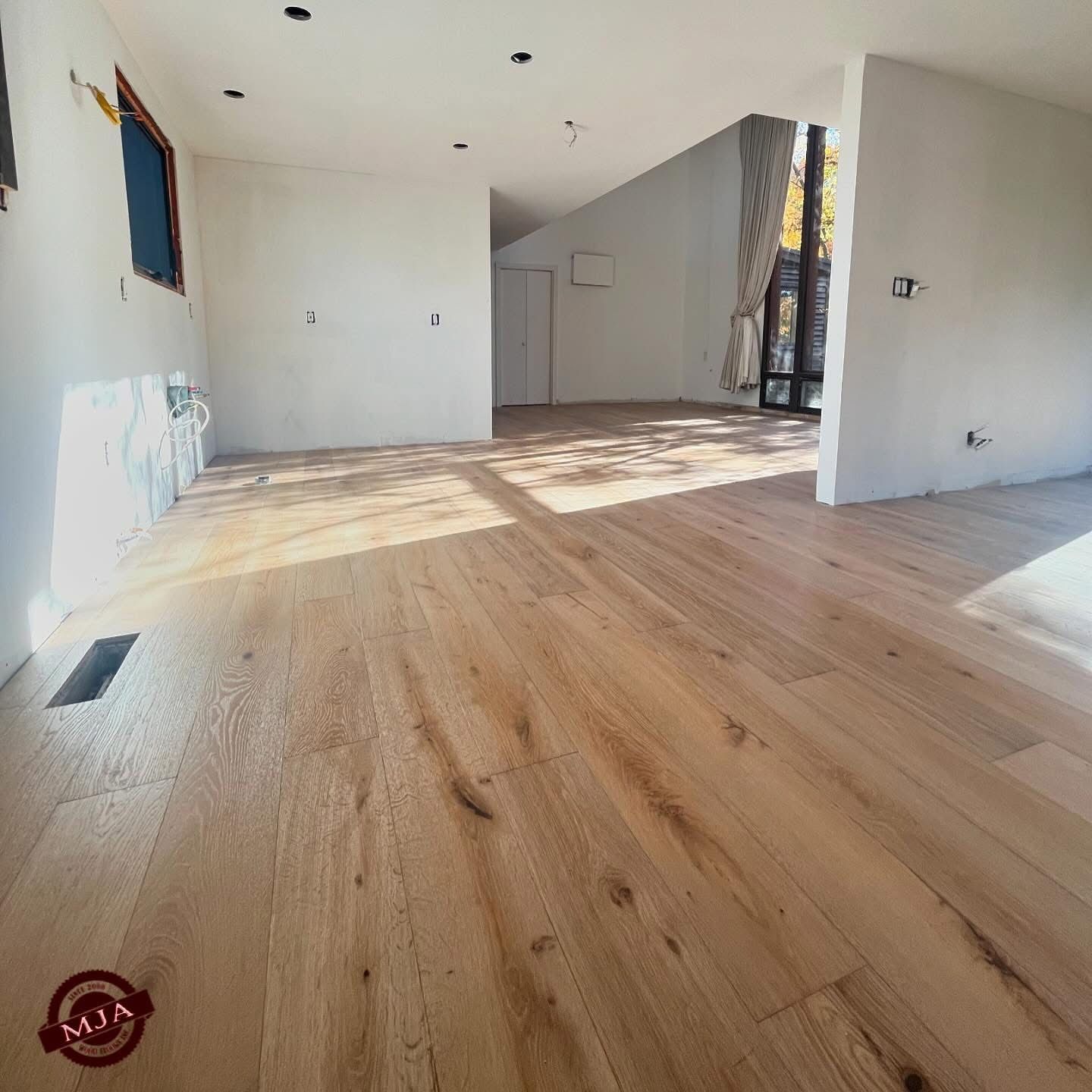 A room with light-colored hardwood floors, white walls, and a large window, under construction.