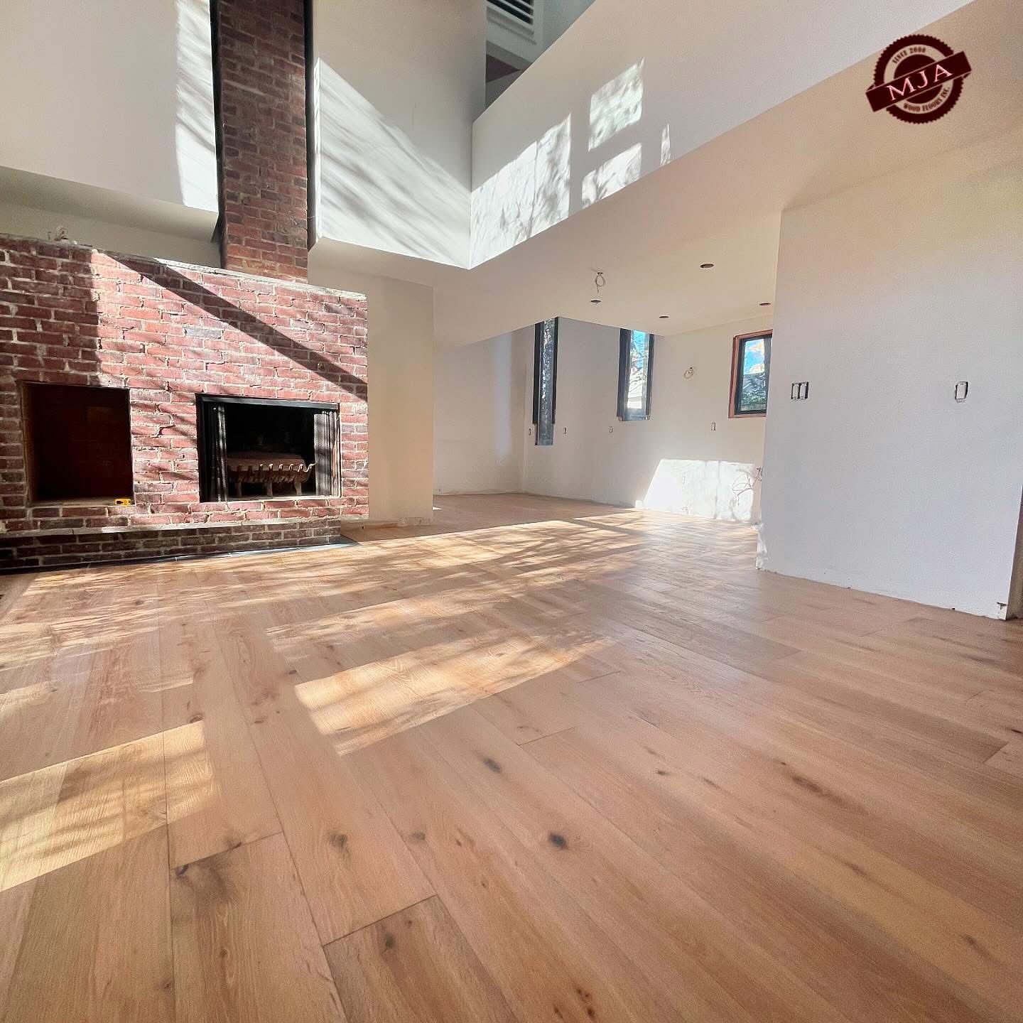A sunlit living room with light hardwood floors, a large brick fireplace, and high ceilings in an open-concept home.