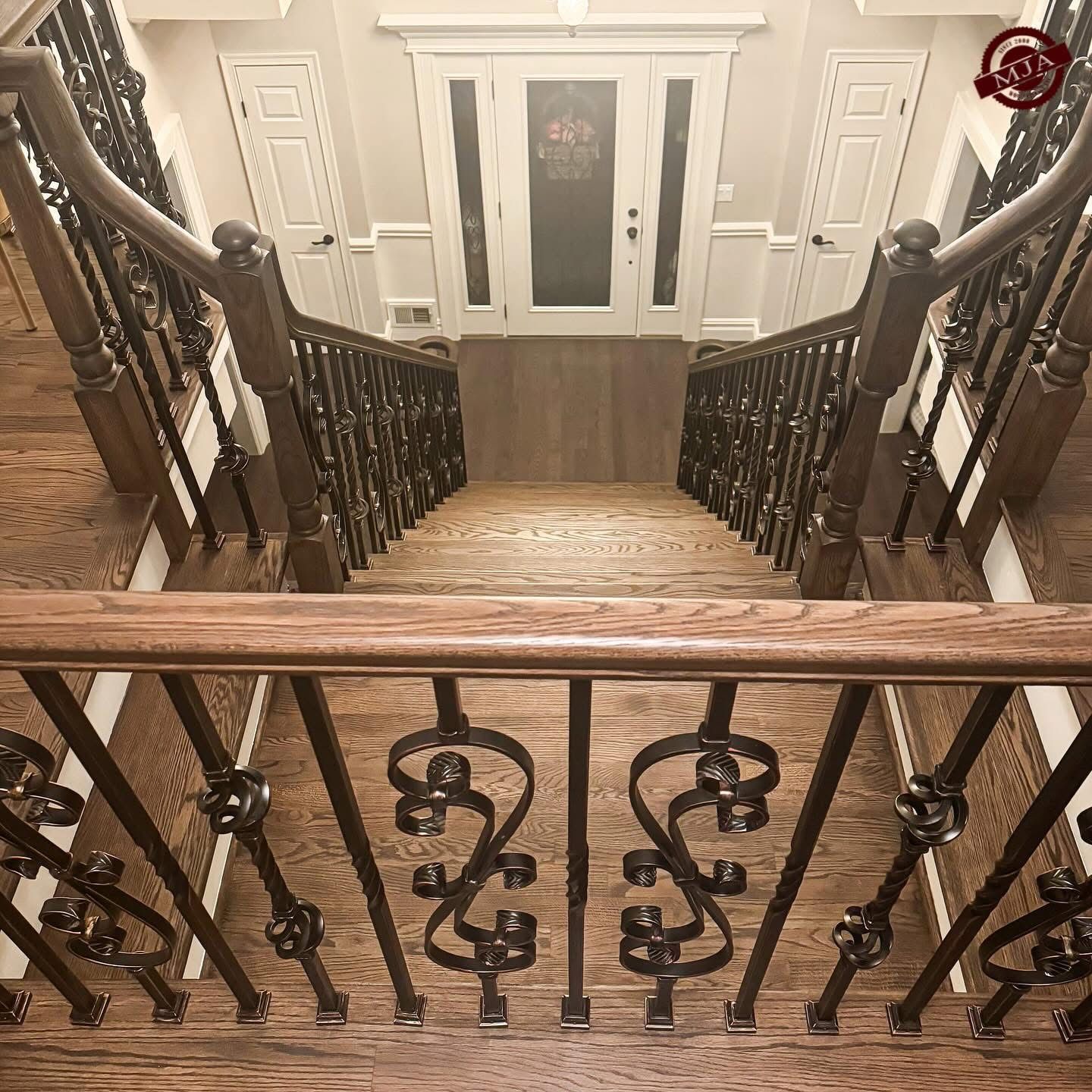 A high-angle view of a wooden staircase with decorative iron railings, leading down to a white double-door entrance.