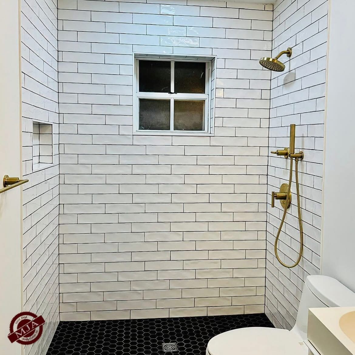 A shower with white textured brick walls, a small central window, black hexagonal floor tiles, and gold-toned fixtures.