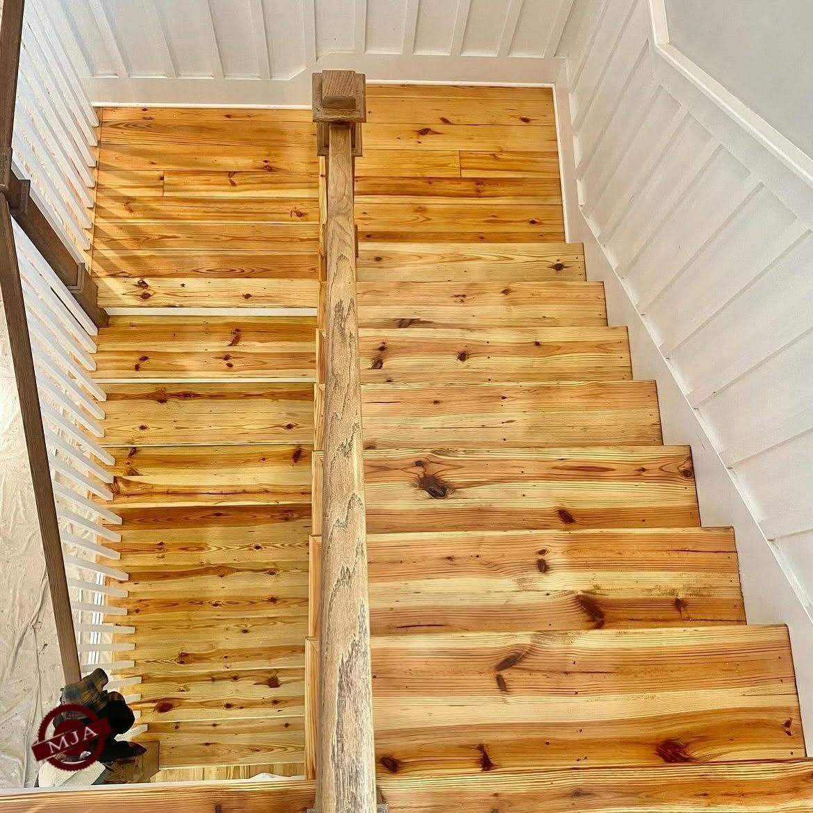 A wooden staircase viewed from above, featuring light-colored planks, a central wooden railing, and white paneled walls.