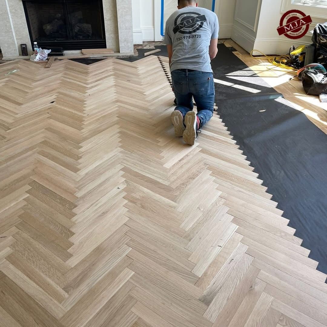 A person kneels while installing light-colored wood floor planks in a herringbone pattern next to black underlayment.