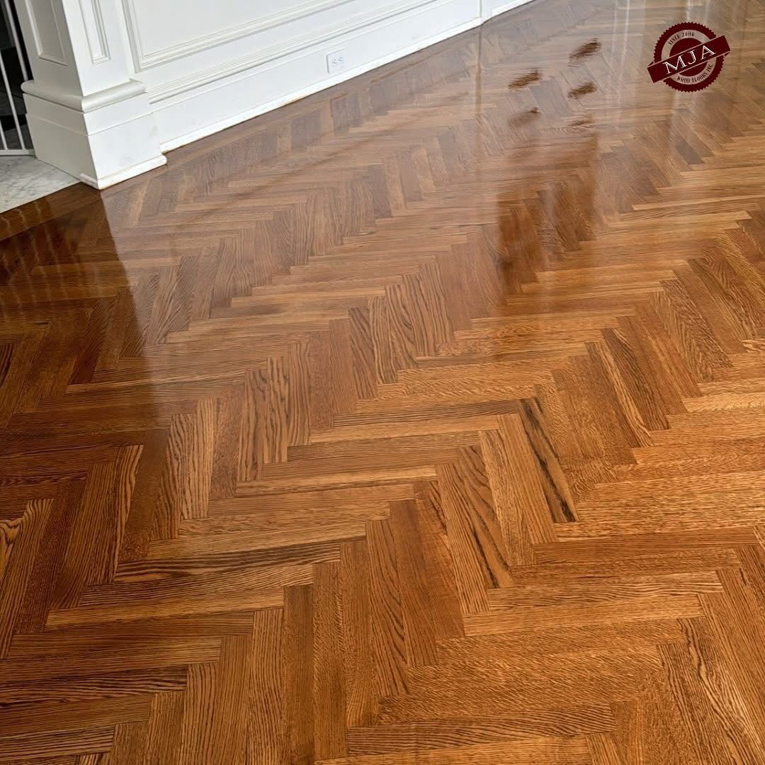 A close-up of a polished brown hardwood floor featuring a herringbone pattern, set next to white baseboards.