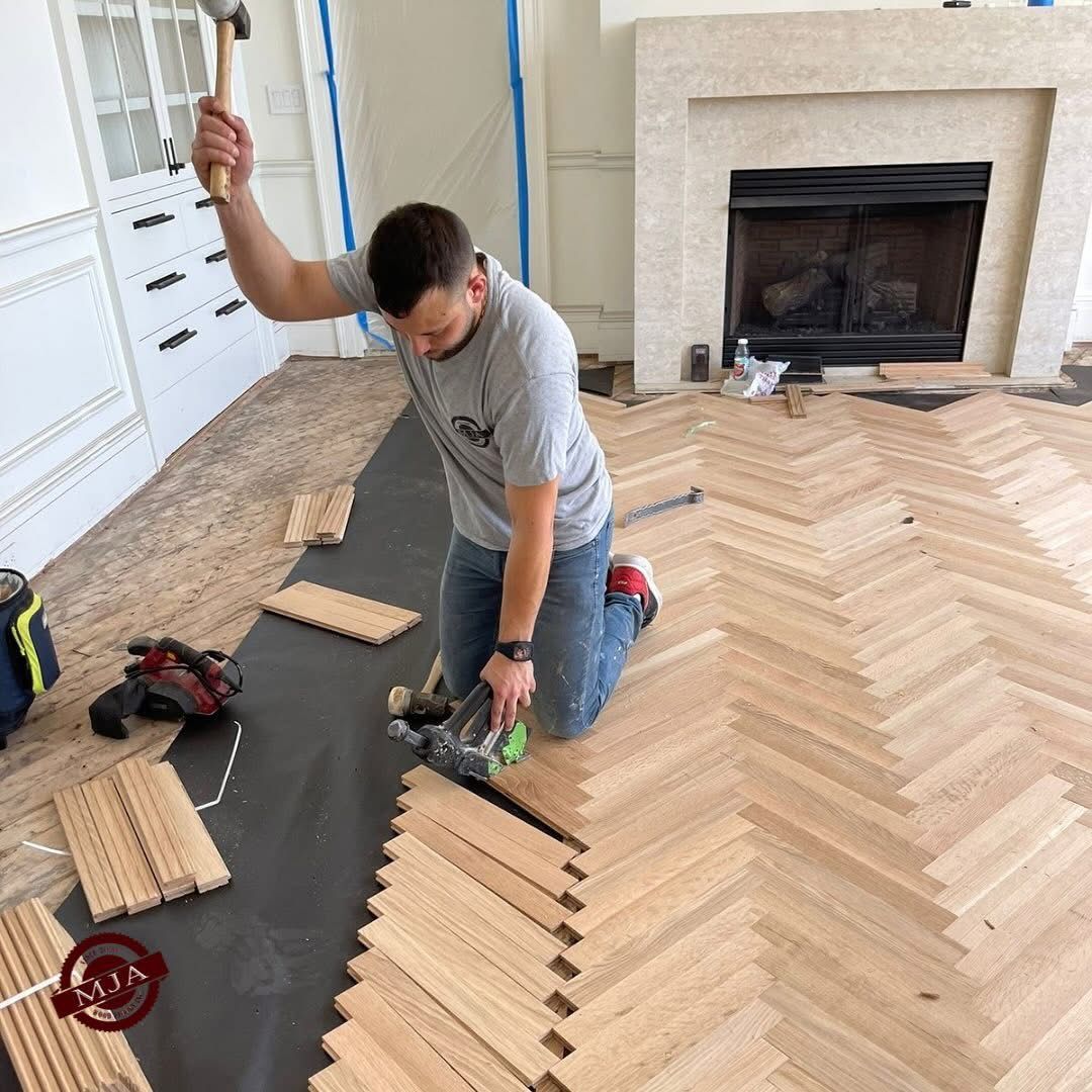 A worker kneels on a floor, installing light-colored wood planks in a herringbone pattern with a nail gun and hammer.