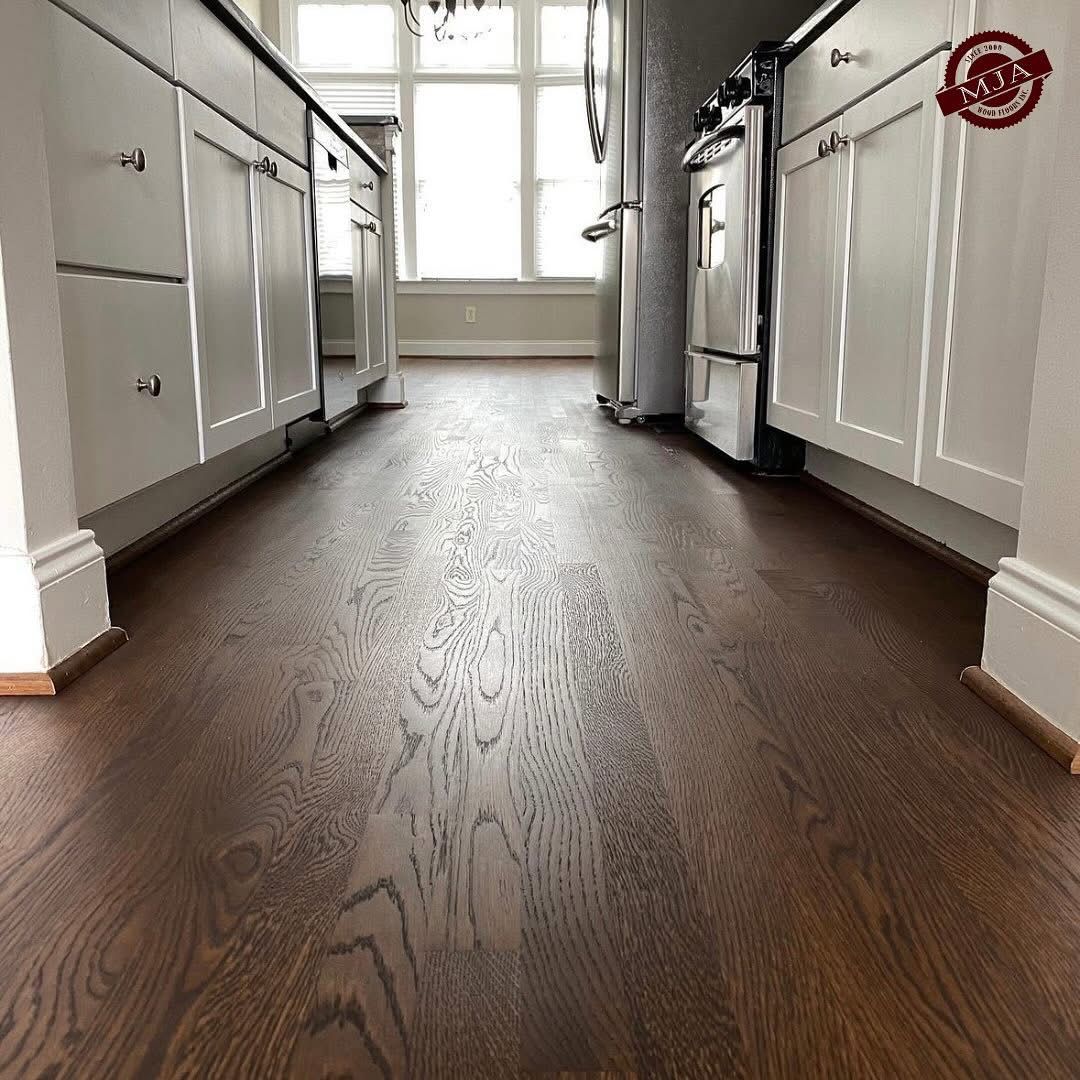 A low-angle view of dark brown wood flooring in a kitchen, flanked by white cabinets and a stainless steel stove.