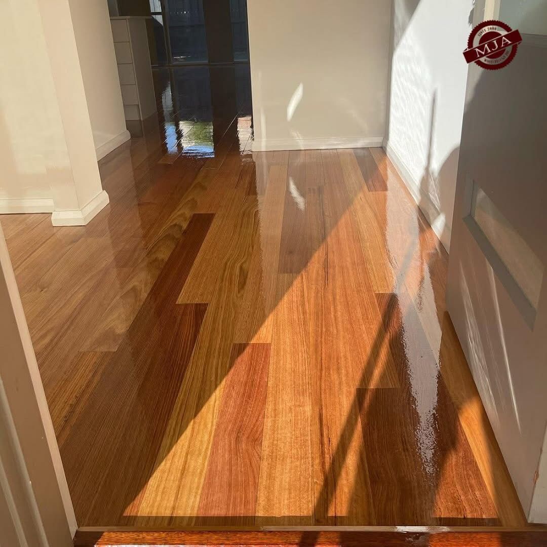 Polished, warm-toned hardwood flooring in a room entryway, illuminated by bright natural sunlight from a nearby door.