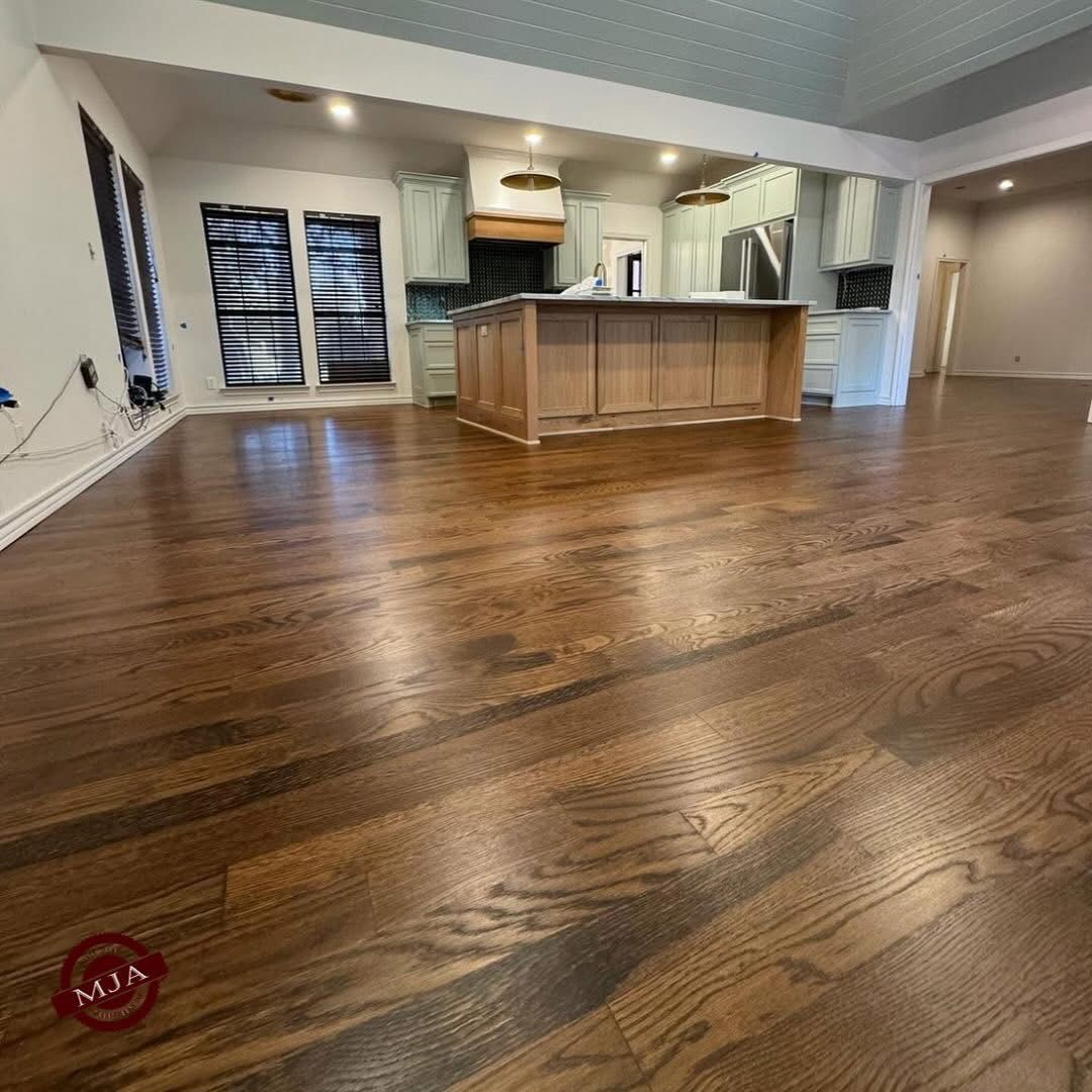A spacious room with newly refinished medium-brown hardwood floors leading to a kitchen with a light wood island.