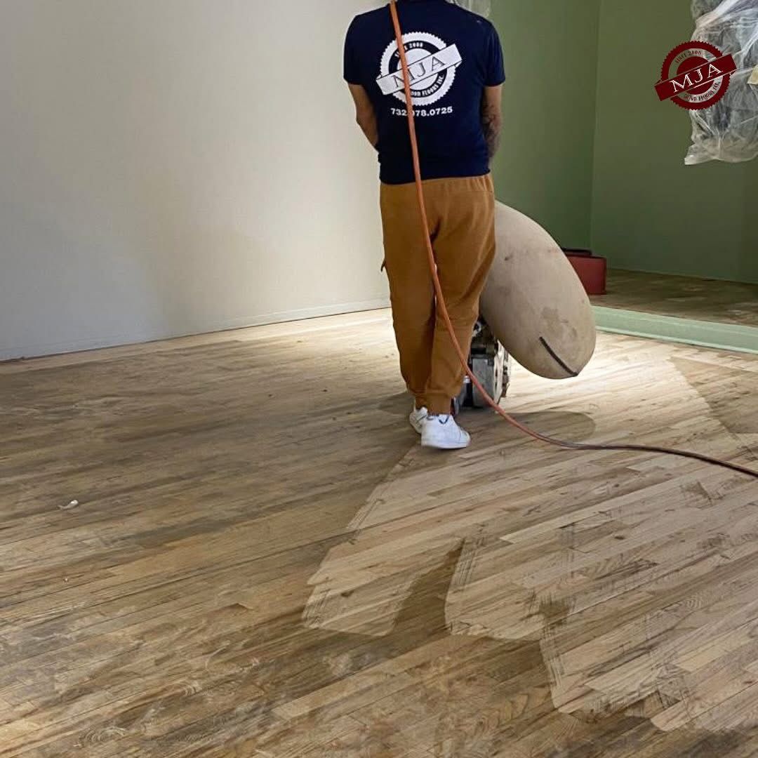 A person in a dark shirt and tan pants uses a floor sander to refinish a wooden floor in a room.