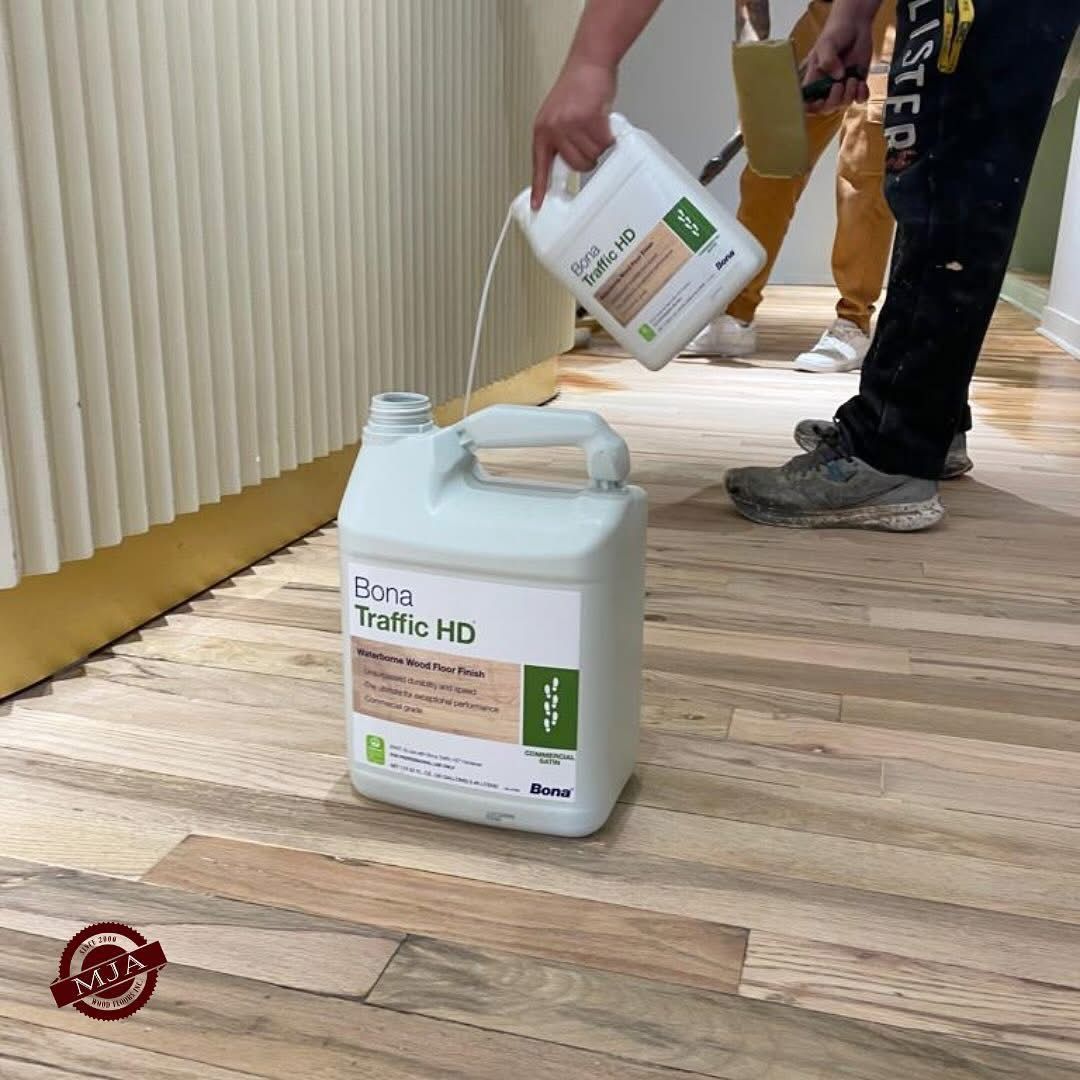 A person pours Bona Traffic HD hardwood floor finish from a smaller container into a larger jug on a wood floor.