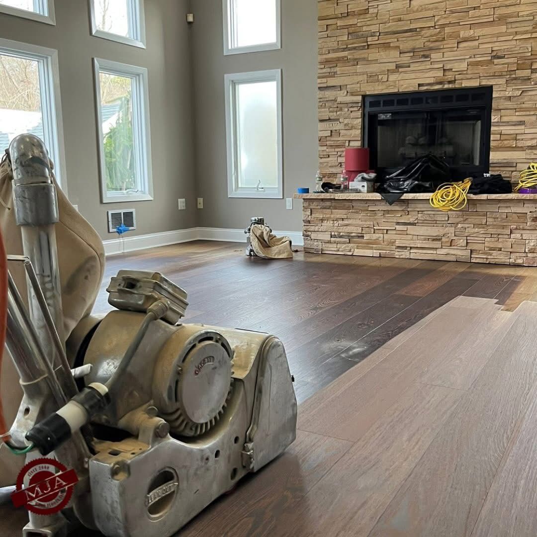A heavy-duty floor sander sits on a partially refinished hardwood floor in a room with a stone fireplace.