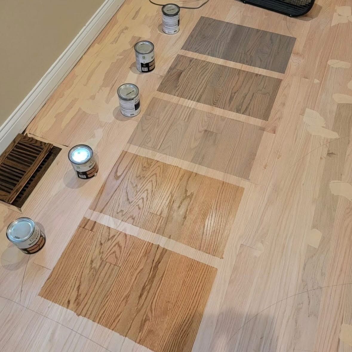 Five rectangular stain samples in shades of gray, brown, and tan are painted onto a light-colored wood floor.