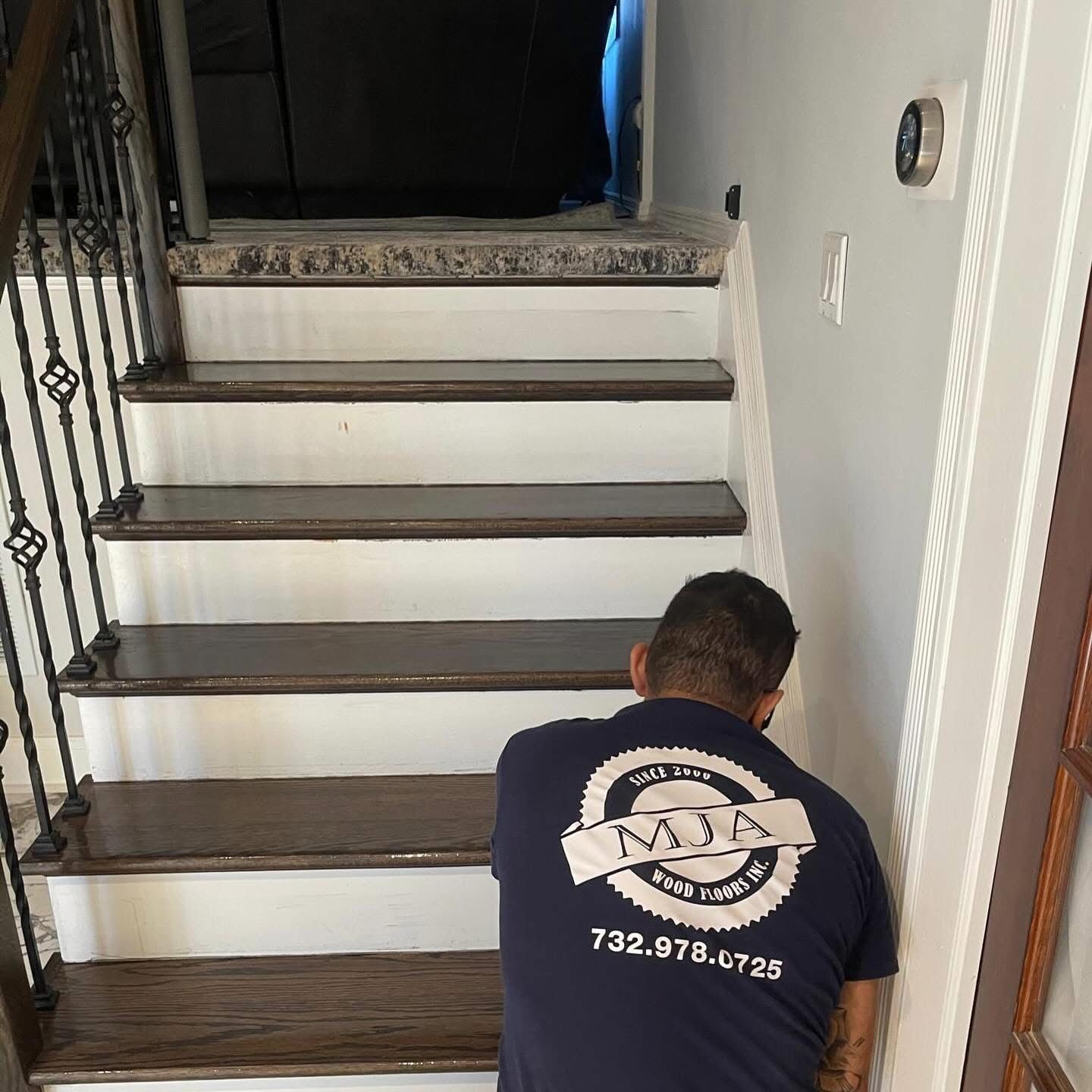 A professional worker in a branded navy shirt works on dark-stained wooden stair treads in a home hallway.