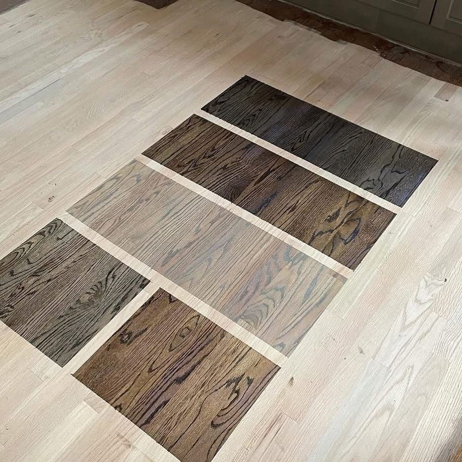 Five rectangular wood stain samples of varying shades ranging from light gray to dark espresso on a light oak floor.