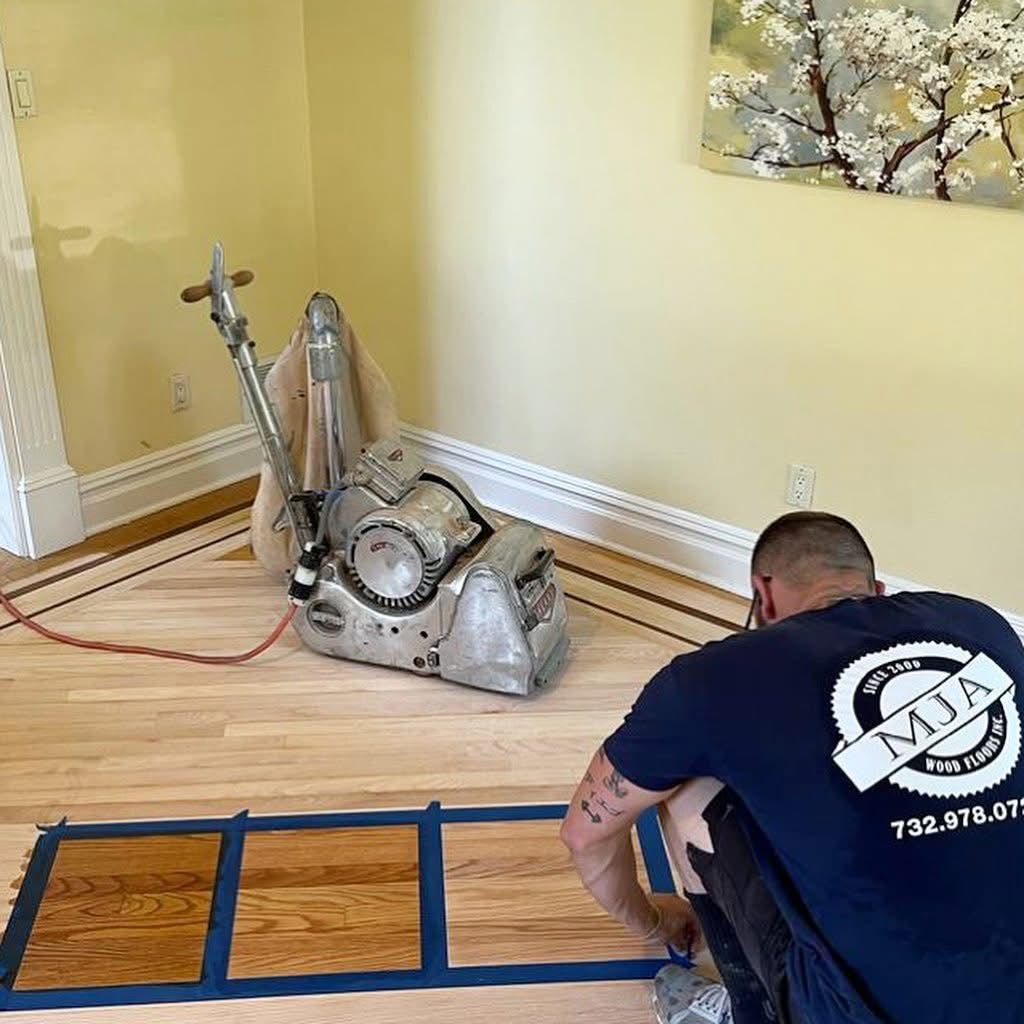 A person in a company shirt applies wood stain samples to a sanded hardwood floor near a large sanding machine.