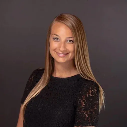 A professional headshot of a smiling person with long, blonde-highlighted brown hair wearing a black lace top.