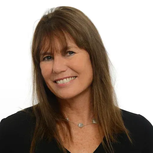 A smiling person with long, shoulder-length brown hair and bangs, wearing a black shirt and a light-colored beaded necklace.