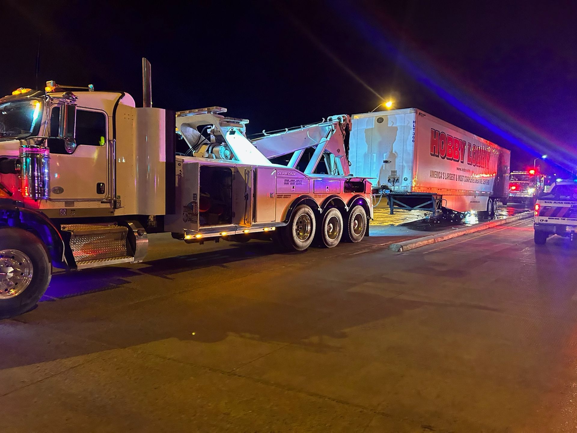 A tow truck is towing a trailer down a street at night.