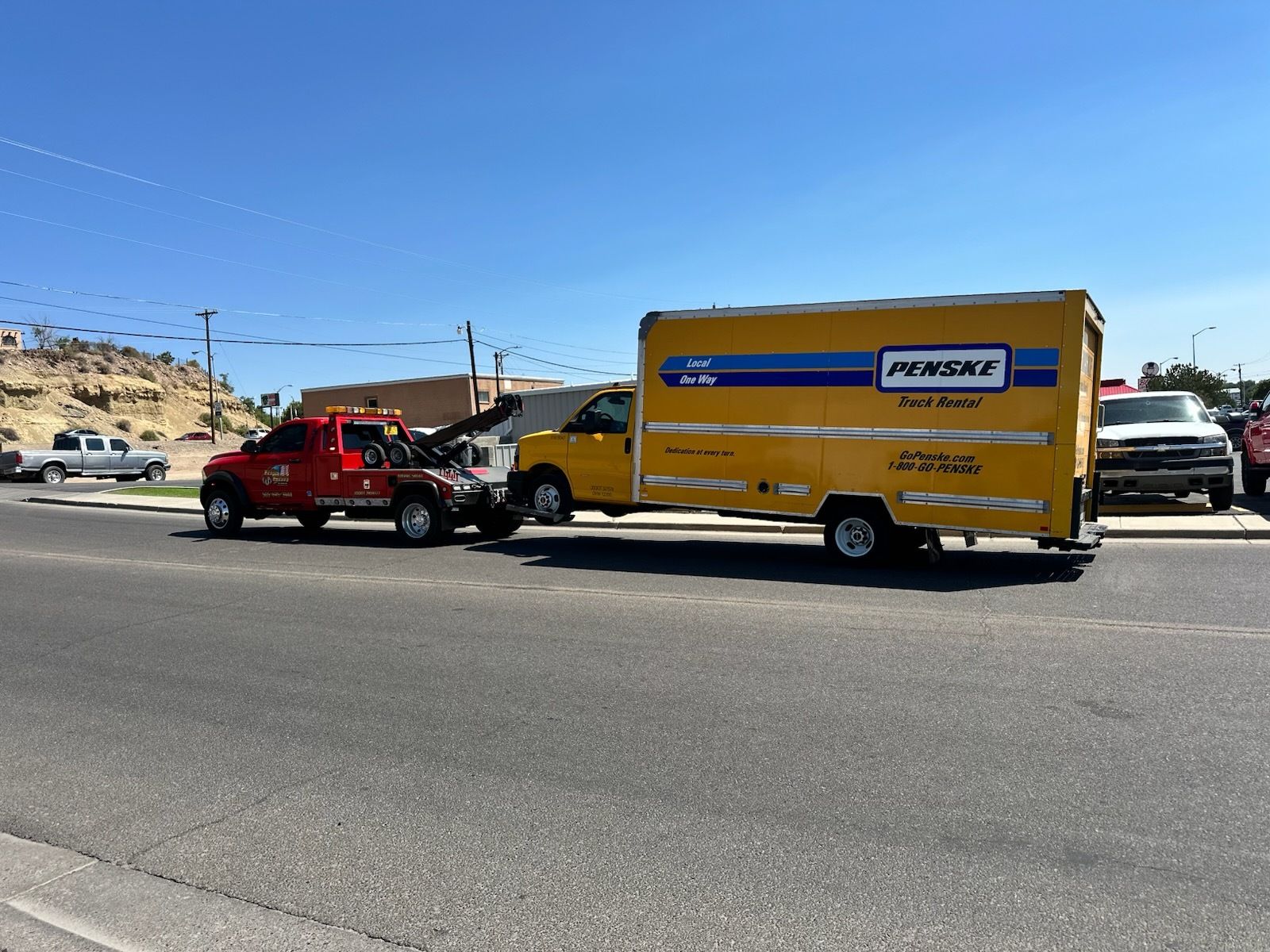 Red tow truck towing a yellow Penske truck on a sunny road.