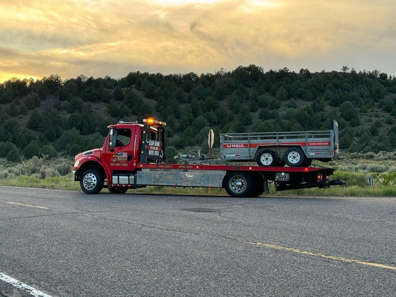 A red tow truck is driving down the road with a trailer attached to it.