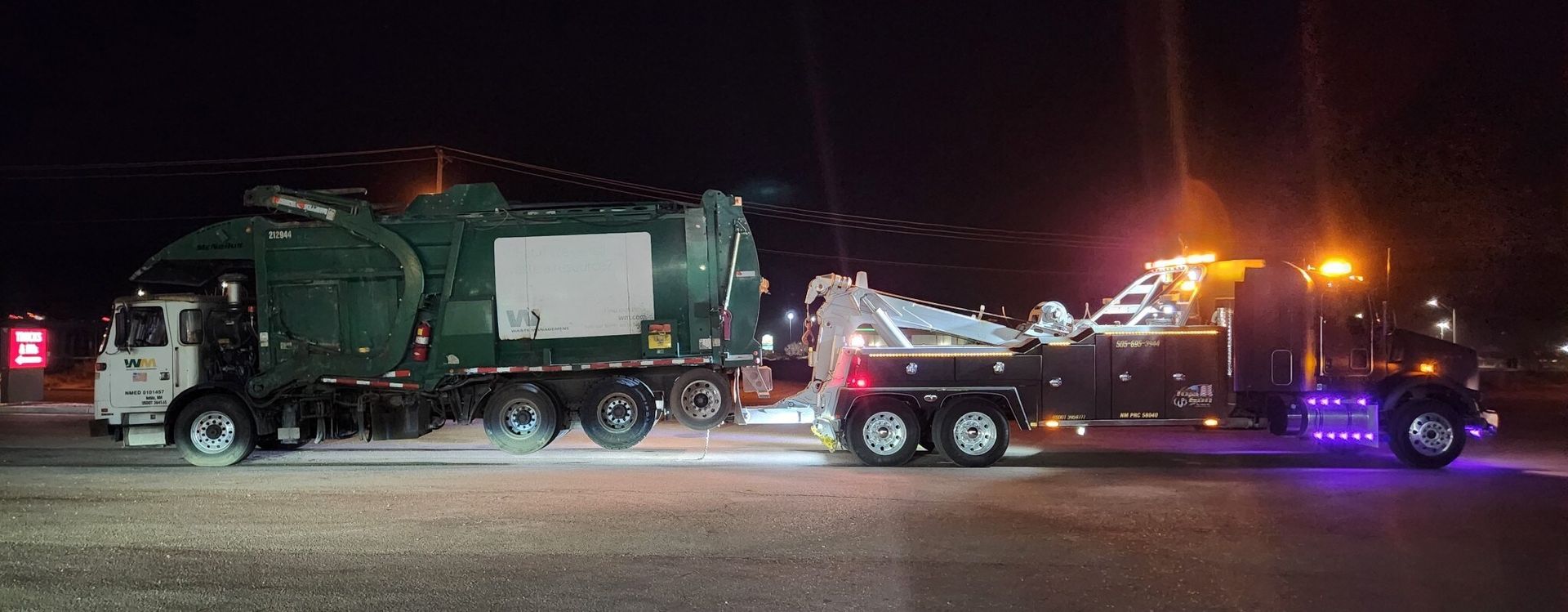 A garbage truck is being towed by a tow truck at night.