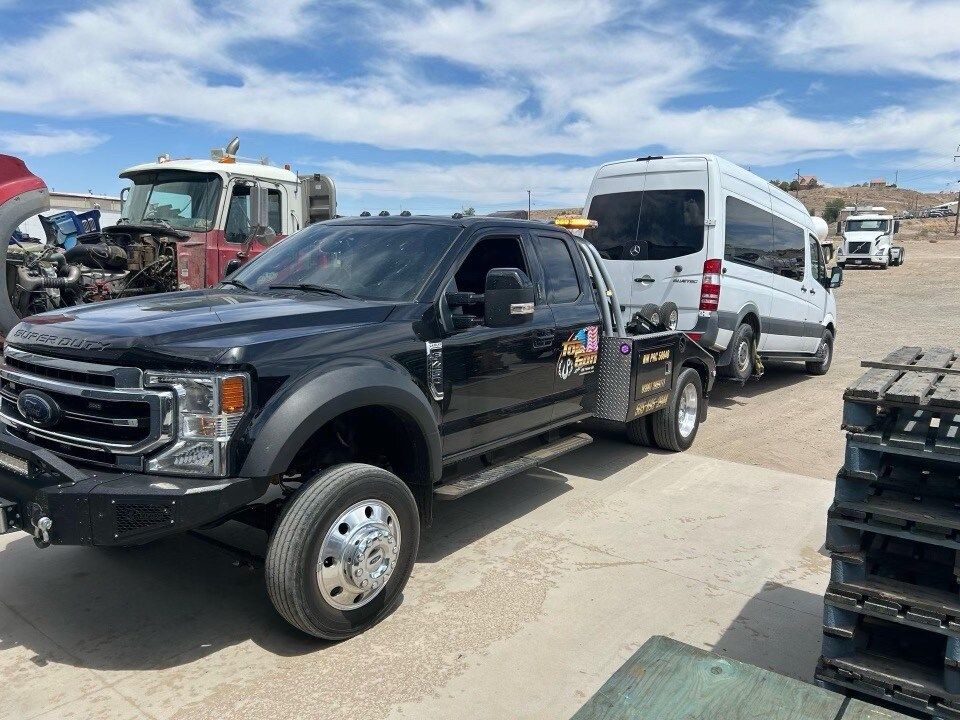 A black truck is towing a white van in a parking lot.