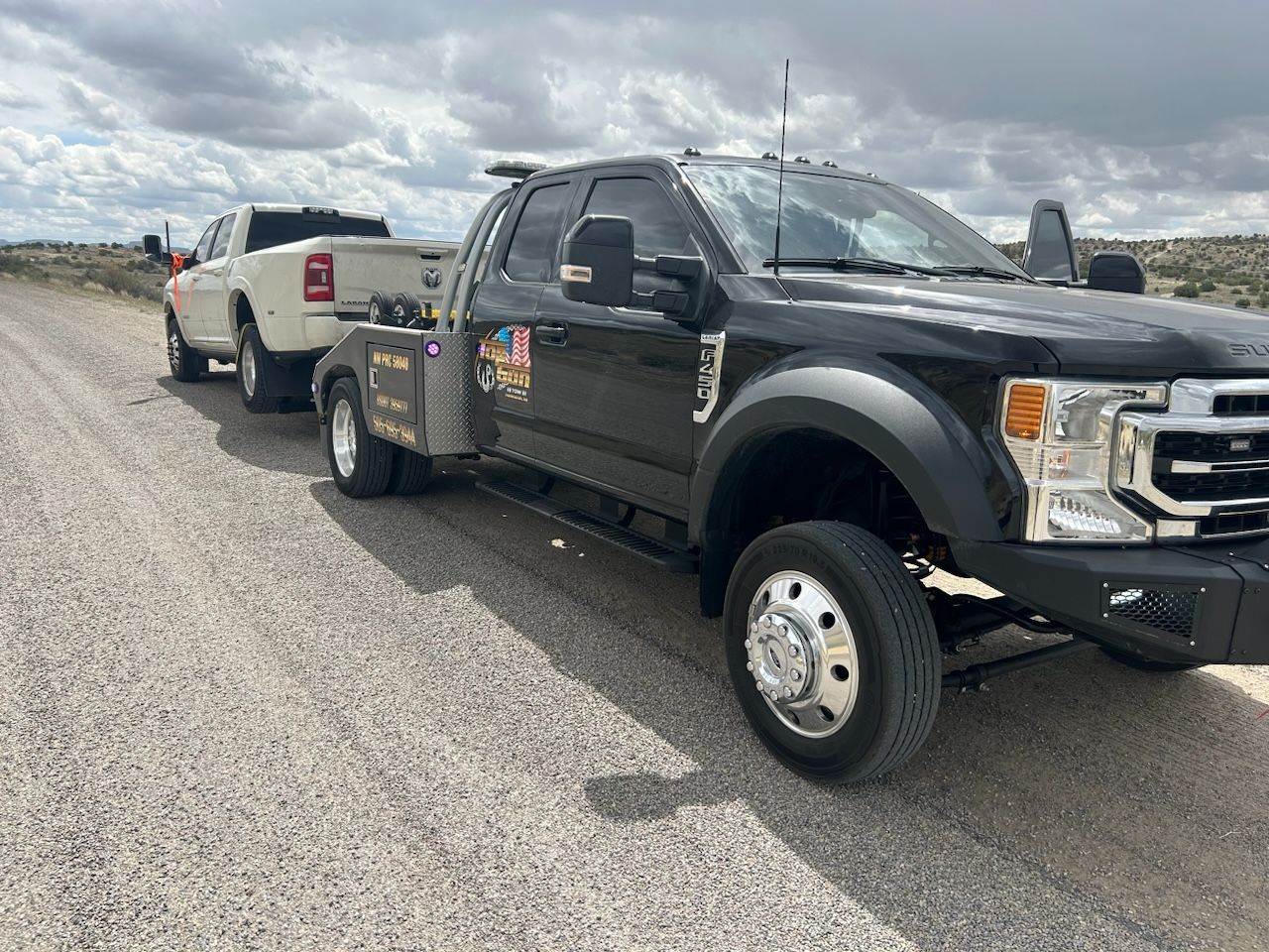 A black truck is towing a white truck on a dirt road.